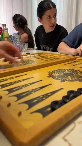 The scene focuses on a backgammon board placed on a table, surrounded by people, drinks, and glasses. The board is yellow with black triangles, with white pieces lined up on one side and black pieces grouped on the other. Several glass bottles and cans of drinks are scattered around the board, along with some drinking glasses. Three people are visible around the table, with one person seated directly across from the camera, appearing to be engaged in conversation. The environment suggests an indoor setting, likely a home or apartment, with natural light coming in through a window behind. The focus of the video seems to be on the backgammon game and the social gathering around it.