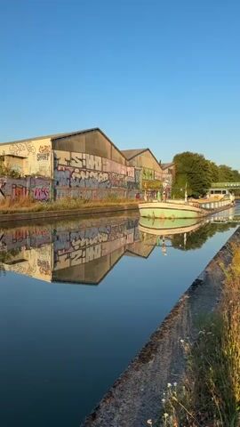A boat named Maritime sails down a canal. The boat is green and white and has a flat deck. The canal is bordered by a concrete walkway on the right and a graffiti-covered building on the left. Trees are visible in the background. The sky is clear and blue. The sun is shining. The water reflects the boat and the building. The scene is peaceful.