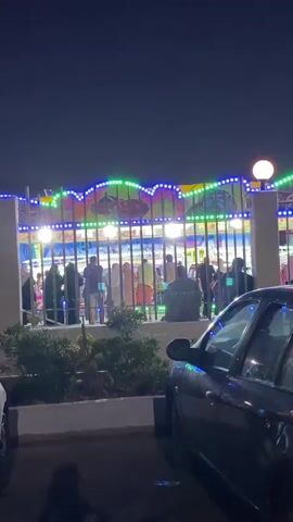 The video shows an amusement park at night. A Ferris wheel is visible in the background, lit with colorful lights. In the foreground, a dark-colored car is parked. Several people are standing behind a fence, appearing to be waiting in line or observing the rides. The atmosphere is festive and lively, with lights illuminating the park.