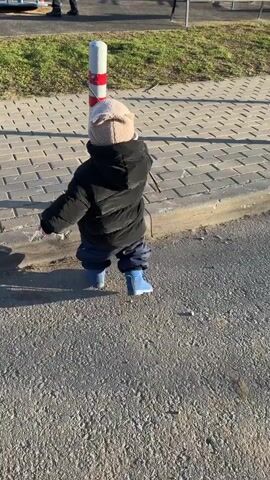 A toddler, viewed from behind, walks on an asphalt path next to a curb and a paved sidewalk. The child wears a black puffy jacket, dark blue pants, light blue boots, and a light-colored beanie hat with small ear-like protrusions. They hold a thin, dark stick in their right hand, occasionally tapping it on the ground or against a white and red striped pole lining the curb.

In the background, beyond the sidewalk and a grassy strip, a children's playground is visible. It features a green slide, yellow benches, and other children playing. Further in the background, multi-story apartment buildings and parked cars can be seen, indicating a residential area. The scene is brightly lit by sunlight, casting long shadows, suggesting either morning or late afternoon. The weather appears clear and cool. The toddler proceeds slowly, exploring their surroundings, at one point stopping to tap a pole before continuing to walk forward. The setting is an outdoor space in Akulovo (Gp Kubinka), Russia.