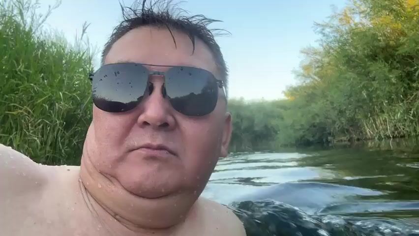 A close-up, over-the-shoulder shot captures a middle-aged man with dark hair, wearing black aviator sunglasses, as he immerses himself in a body of water. Water droplets are visible on his face and shoulders, suggesting he is actively swimming or wading. The water appears to be a river or stream, with visible ripples and movement. Tall, green grass and foliage line the banks of the water body, creating a natural, outdoor setting. The sky is visible in the upper portion of the frame, indicating it is likely daytime. The man's expression is neutral, and he is looking directly at the camera. The camera appears to be held by the man himself, creating a selfie-style perspective.