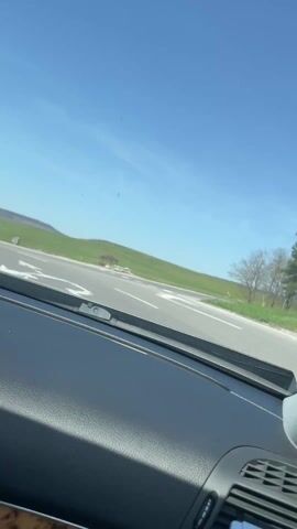 The video is shot from the driver's seat of a car approaching a roundabout on a sunny day.  The sky is clear and blue. The countryside is green with rolling hills and some trees.  A minivan is visible passing to the left. The interior of the car is visible in the foreground, showing the dashboard and part of the center console. There is dance music playing.