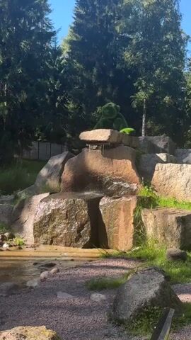 A stone waterfall is in the foreground with a green moss-covered bear figurine sitting on top of the waterfall. The bear is facing to the left. Water is flowing over the rocks and into a shallow pool at the bottom. The background consists of trees and a blue sky.