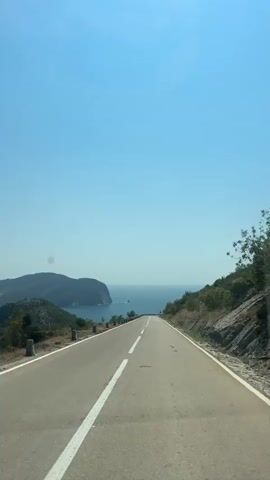 The video shows a point-of-view shot from a vehicle traveling along a winding road with the ocean visible in the distance. The road is paved and has a white dashed line marking the center. On the right side of the road, there is a steep, vegetated slope with rocks. On the left side, there are some rocky outcrops and sparse vegetation, with a railing visible along the edge of the road. In the distance, hills and the sea can be seen under a clear blue sky. A speed limit sign indicating 40 is visible on the right side of the road. The sun appears to be shining brightly.
