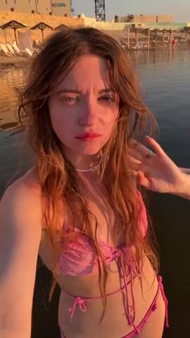 A woman with long, wavy, light brown hair, wearing a pink polka dot bikini, stands in shallow water near a beach. The water reflects the warm golden light of the setting sun. In the background, there are beach chairs with umbrellas and buildings. The woman makes various facial expressions and poses for the camera, sometimes touching her hair or arm. The video is a close-up selfie-style recording.