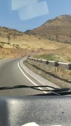 The video shows a view from inside a vehicle, looking out the windshield and side windows. The vehicle appears to be driving on a paved road that curves to the left, flanked by a metal guardrail with alternating black and white reflective sections. The landscape is arid and mountainous, with dry, yellowish-brown vegetation covering the hillsides. The sky is clear and blue. In the distance, more rugged mountains are visible. The camera pans slightly to the right, revealing more of the dry, hilly terrain. A side mirror is visible on the right side of the vehicle, reflecting the road and surrounding environment, showing another vehicle behind. The video captures a sunny day in a natural, undeveloped area.