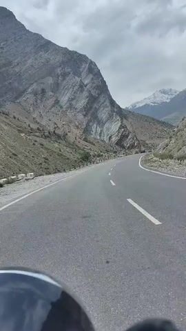 The video captures a POV shot from a vehicle, likely a motorcycle, driving on a paved road that curves through a mountainous landscape. The road has clear white lane markings. On the left, a steep, rocky mountain with visible strata rises dramatically. On the right, the terrain slopes downwards into a valley, with more mountains visible in the distance, some of which are snow-capped. The sky is overcast with heavy clouds, suggesting a cool or potentially rainy day. The foreground is dominated by the helmet or windshield of the vehicle the video is being filmed from. No people are clearly visible, and the overall impression is one of a solitary journey through a rugged, remote environment.