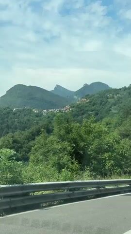 The video shows a view from a moving vehicle on a road. The road has a white line on the right side and a metal guardrail on the left. The surrounding environment is mountainous and green, with trees and grass. In the distance, there are buildings on the side of a hill and a clear sky with some clouds.