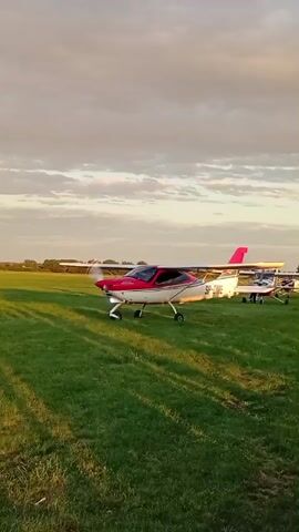 The video shows a small white and pink airplane with the registration SP-DME on a grassy field during golden hour. The airplane is stationary, with its propeller visible and appearing to be intact. The sun is low in the sky, casting long shadows across the grass. The sky is partly cloudy, with a mix of light and dark clouds. In the background, there are trees and what appears to be another aircraft parked on the field. The overall atmosphere is serene and peaceful.