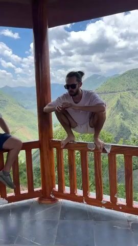 A medium shot shows a man balancing in a squat on a railing. The man wears sunglasses, a white tee-shirt, and white shorts. He has a beard and a man-bun. He has his hands together near his chin, and he smiles. His feet are bare. He is on a wooden balcony with a wood railing. The railing is worn, and the paint is peeling. Behind the railing is a panoramic view of lush green mountains under a sky with puffy clouds.