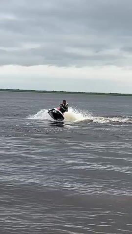 A person is riding a jet ski on a body of water. The jet ski is black and red. The person is wearing a black and white life vest and dark shorts. The person is leaning to the left, causing the jet ski to spray water. The sky is cloudy. The water is dark and choppy. In the background, there is a distant horizon with a green line, likely representing land. The video captures the motion of the jet ski and the spray of water.