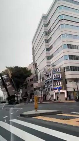 The video shows a street scene in what appears to be a city in Japan. The camera appears to be in a vehicle moving through an intersection. There are several buildings of varying heights and architectural styles, including a tall, modern office building with a multi-story advertisement on the side of it. There are a few pedestrians visible on the sidewalks. Traffic lights are seen changing from red to green. The day is overcast and the streets are wet, possibly from rain. A small white truck is visible stopped at the intersection.