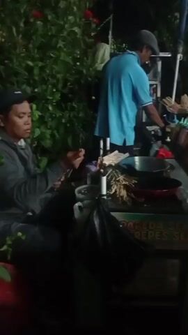 Two individuals are engaged in food preparation at an outdoor night market stall. In the foreground, a person wearing a grey hooded sweatshirt and a black baseball cap is seated and appears to be preparing skewers. To the right, another individual in a blue t-shirt and grey baseball cap stands and is actively cooking on a grill or flat-top surface. A third person, a woman wearing a dark t-shirt and shorts, stands to the far right and is holding a skewer. The stall is decorated with signage that reads "PAREDA SELLA CREPES SELLA." Various ingredients and cooking utensils are visible on the counter, including bottles of sauces, skewers, and pans. The background is filled with lush green foliage, suggesting an outdoor setting. The lighting is dim, indicating it is nighttime.