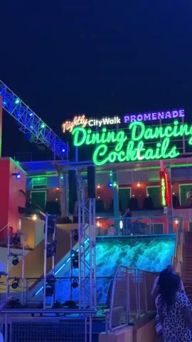 The video captures a lively night scene at CityWalk Promenade. A brightly lit sign announces "Nightly CityWalk PROMENADE Dining Dancing Cocktails" in green and yellow neon. Below the sign is a multi-level building with outdoor seating areas, visible through the railings.  A waterfall display with blue lighting flows down the front of the structure. The camera pans across the scene, showing a large screen with colorful projected images. People can be seen walking and standing near the base of the building, including a man in a white shirt and cap using his phone. Lighting rigs and other equipment suggest an active entertainment venue.