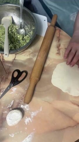 Here is a description of the video:

A person is preparing dough on a patterned surface that resembles marble. The person folds the dough in half, then in half again, forming a quarter circle. There is a metal bowl to the left filled with a green mixture. A rolling pin is centered in the scene, and a pair of scissors are also visible. A ball of dough is in the lower left corner, dusted with flour.