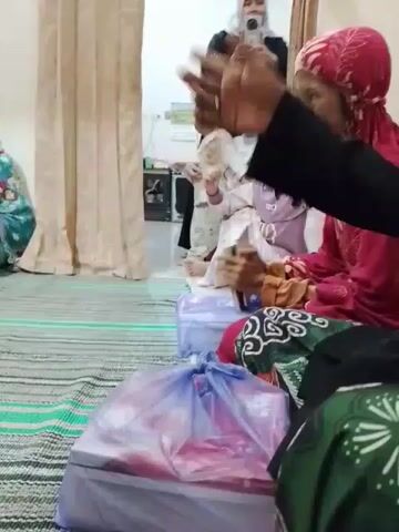 The video shows a group of people sitting on a carpeted floor in a room. Several children are visible, some wearing hijabs. A woman in a pink hijab is holding a smartphone. A man wearing a black jacket and a traditional Muslim cap is also present. In the foreground, there's a gift wrapped in a lilac-colored plastic bag. The people seem to be engaged in a gathering, possibly a celebration or a family event. The room is simply furnished with white walls and minimal decor.