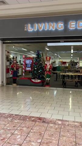The video pans from a storefront with Christmas decorations, including nutcracker figures, a Christmas tree, and a Santa countdown timer, to an empty shopping mall corridor. The storefront displays the sign "THE LIVING ROOM." Several shops and an exit sign are visible in the background. The mall features tiled floors and natural light from skylights. There are no people clearly visible in the mall, except for a distant figure walking away from the camera. The overall atmosphere is quiet and empty.