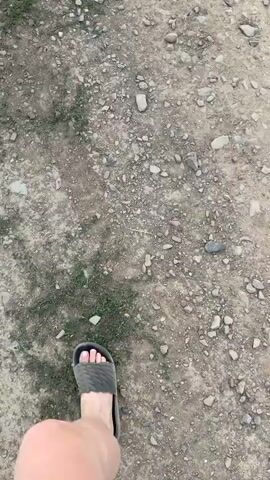 This video begins with a shot from behind three individuals walking on a dirt path. The person in the middle wears black shorts and white socks with athletic shoes. To their right, a person in light-colored pants and a patterned shirt walks away from the camera. To the left, a person in dark pants and white sneakers is partially visible. The path is flanked by grass and a field of dry, golden grain on the right. The camera pans to show the person in the patterned shirt walking past the grain field, looking towards a cloudy sky and distant mountains. The scene then shifts to a close-up of a woman with dark hair, wearing a white tank top, with a blurry background of a field and sky. The final shot shows the dirt path next to the grain field.