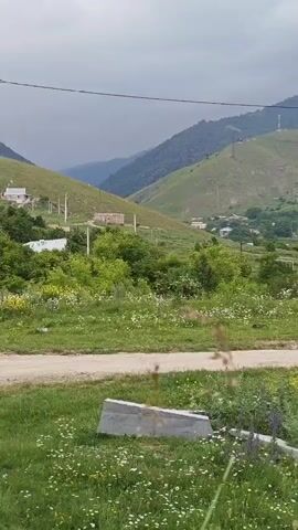 This media file depicts a scenic rural landscape in Gugark, Armenia. The scene is dominated by rolling green hills, some dotted with scattered houses and what appear to be communication towers on the highest peak. A dirt road winds through the foreground, bordered by lush green grass and wildflowers, primarily white daisies and some purple lupine. In the mid-ground, a cluster of houses forms a small village, nestled amongst trees and more verdant hills. The overall impression is one of a peaceful, somewhat remote, countryside setting.

The time of day appears to be daytime, likely overcast or with a muted sun, as the light is soft and diffused, casting no harsh shadows. The weather seems to be temperate, with abundant greenery suggesting a mild season, possibly late spring or summer. The presence of a utility pole on the left and a pile of concrete slabs in the foreground suggests some level of human habitation and perhaps ongoing construction or infrastructure work in the immediate vicinity, contrasting with the natural beauty of the broader landscape.

There are no people visible in the media file. The focus is entirely on the natural environment and the small village. The absence of people and the tranquil scenery evoke a sense of quietude and simplicity. The architecture of the houses appears typical of rural Armenia, with pitched roofs. The location cues, combined with the visual elements, strongly suggest a rural Armenian setting.

No visible text is present in the media file. The overall mood is serene and picturesque, highlighting the natural beauty of the Armenian countryside.