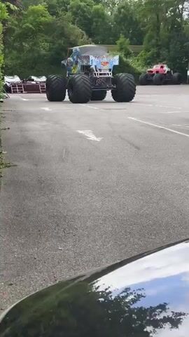 A monster truck with a blue and white design is parked on an asphalt lot. Another red monster truck is visible in the background. A person is standing in front of the blue and white truck. The ground is paved with asphalt and marked with a white arrow. There are trees and foliage surrounding the lot. The person walks away from the blue and white truck.