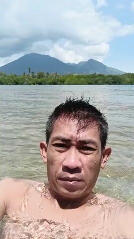 The video shows a man floating in a body of water, presumably a river or lake, on a sunny day. He is seen from the chest up, facing the camera, with water touching his neck. In the background, there is a shoreline with trees, followed by landmasses shaped like hills or mountains. The sky is blue with scattered white clouds. The camera perspective suggests the person is holding the camera in front of them while in the water.