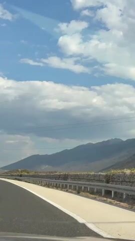 The video shows a point-of-view shot from inside a moving vehicle on a highway. The road ahead curves to the left. There are white lane markings on the asphalt road. On the right side of the road, there is a guardrail and a stone wall. Beyond the wall, there are sparse bushes and rocky, mountainous terrain. The sky is blue with scattered white and gray clouds. In the distance, a red vehicle is also traveling on the highway ahead. In the lower part of the frame, the dashboard and windshield wipers of the vehicle are visible.