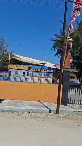 The video shows a roadside scene with a blue sky and some trees in the background. The road is unpaved with some gravel and dirt. There are several poles with electrical wires running across the scene. A building with an orange and white facade is visible, along with a fence. A flag with some writing on it is attached to a pole. The camera pans across the scene, showing more of the building and the road, and then a truck and a motorcycle are seen driving on the paved road in the distance. The overall impression is of a rural or semi-urban environment on a sunny day.