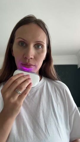 A woman in a white t-shirt is standing in front of a camera, holding a white device with a curved top that emits a pinkish-purple light. She moves the device along her jawline and cheek, seemingly using it for skincare. Her brown hair is loose, and she wears a ring on her left hand. In the background, a dark-colored wall and a doorframe are visible, indicating an indoor setting. There is ambient noise, including speech, in the background, possibly from a child.