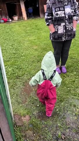 A person in a light green hooded outfit and dark pink overalls, with purple boots, is walking in a grassy area with puddles. The child is facing away from the camera, and has their back to the viewer. Behind them, a person wearing a black and white patterned jacket and black pants with purple boots is standing. The ground is covered in green grass and several puddles of water. There is a wooden structure in the background. The video is 5 seconds long and appears to be filmed outdoors.