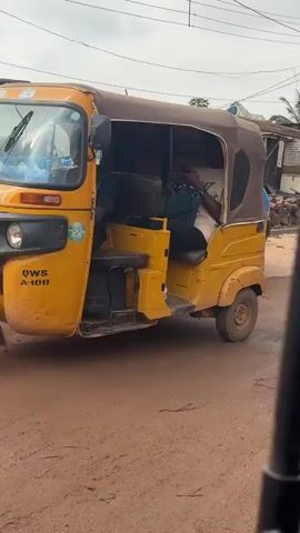The video depicts a street scene with various vehicles and people. In the foreground, a black motorcycle is moving, carrying two individuals. The driver is wearing a dark jacket and a cap, while the passenger behind him is wearing a green shirt. Further down the road, a red auto-rickshaw is visible, with two people inside. The driver appears to be wearing a brown shirt, and the passenger is in a green shirt. The auto-rickshaw is on a dirt road, and in the background, there are market stalls with various goods, including sacks of produce and piles of wood. Another yellow auto-rickshaw is seen in the distance. The environment appears to be an outdoor market or a busy street in a town. The sky is overcast.