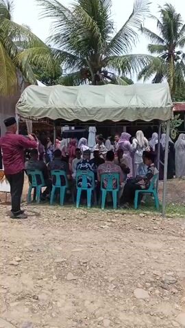The video shows an outdoor gathering of people, possibly a community event or a memorial service. The scene is set under a tent, suggesting shade from the sun. Several individuals are seated on chairs, while others stand or move around. Some people are dressed in traditional attire, indicating a cultural or religious context. In the foreground, a man in a maroon shirt stands and appears to be taking a photo or recording with his phone. There are palm trees and vegetation visible in the background, suggesting a tropical or sub-tropical environment. The ground is unpaved and appears to be dirt or gravel. There are also some black drapes visible, which might indicate a somber occasion. The overall atmosphere seems calm and organized.