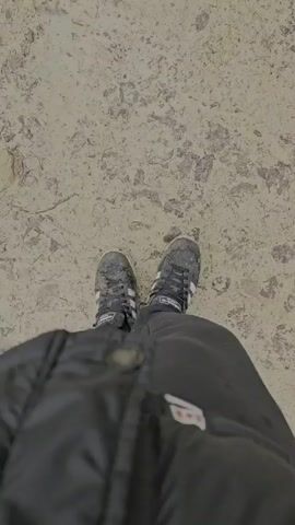 A person wearing Adidas shoes and a black puffer jacket walks on a muddy path. The camera is directed at the person's feet as they walk. The person is walking with a gray dog on the path. The path is surrounded by trees.