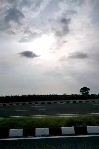 The video shows a view from a moving vehicle on a highway. The sky is cloudy with the sun peeking through. There are tall grasses along the side of the road, and a few trees in the distance. The road has white lines marking the lanes. In the last few frames, a person is seen walking away from the camera on the side of the road. There is also a white car parked on the left side of the road.