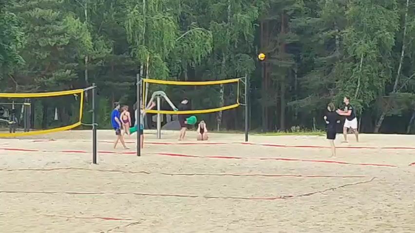 This video depicts a sandy beach volleyball court surrounded by a lush green forest. The scene is set during the daytime, likely late afternoon or early evening, judging by the soft light and the absence of harsh shadows. The weather appears to be warm and pleasant, conducive to outdoor recreation.

Several individuals are engaged in a game of volleyball. In the foreground, a person in a blue shirt is in motion, seemingly about to serve or hit the ball. Nearby, another person in a light-colored top and blue shorts stands ready. Across the net, a person in a black top and shorts is positioned, while a person in a dark top and shorts is in the background. Another individual in a black outfit is standing to the right, observing the game. One person in a black top and green shorts is kneeling on the sand near the net, possibly waiting for play to resume or in a resting pose.

The volleyball court has yellow nets and red boundary lines marking the sand. In the background, there is playground equipment, including swings, adding to the recreational atmosphere of the location. The forest provides a natural backdrop, with tall trees obscuring any distant structures. There is no visible text in the video. The overall mood is one of casual, energetic recreation on a summer day.