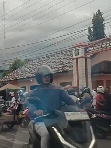 The video depicts a busy street scene with numerous motorcycles and a few cars. The sky is overcast. In the foreground, a black car is visible, followed by several motorcycles. One motorcycle carries three people, all wearing head coverings. Another motorcycle has a driver wearing a brown head covering and light-colored pants. Behind this motorcycle is a flatbed truck. Several other motorcycles and cars are in the background, suggesting traffic congestion. Buildings with tiled roofs are visible along the street. The overall impression is of a crowded urban environment with significant motorcycle usage.