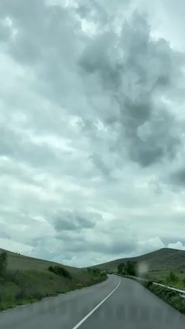 The video depicts a road trip on a gray, overcast day. The camera appears to be inside a vehicle, looking out through the windshield. The road is asphalt, with a white dividing line. On either side of the road, the landscape consists of green, grassy hills. A metal guardrail runs along the side of the road. The sky is dominated by thick, gray clouds, with very little blue visible.