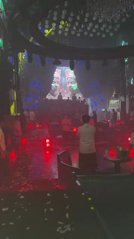 The video shows a nightclub with staff moving around preparing for an event. The club is dimly lit with red accents and confetti scattered on the floor. A DJ booth is visible on stage, with a large screen displaying animated visuals in the background. Yellow spotlights illuminate the stage area. The club appears mostly empty, with only staff members present.