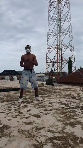A young, shirtless man with a pair of white goggles stands on a concrete rooftop. He wears denim shorts with a repeating black and white pattern and white sneakers. The man is positioned in front of a tall, red and white metal tower, possibly a telecommunications tower, which dominates the background. To the left of the tower, houses with dark roofs are visible, and a single tall green tree stands near the tower. The ground of the rooftop is uneven and textured, with patches of dirt and what appear to be scattered rocks. The sky is overcast with grey clouds, suggesting a possibly humid or rainy day. The man's posture and the way he holds his arms, with one hand on his hip and the other extended slightly, suggest he is posing or perhaps engaged in a dance or martial arts stance. The overall scene appears to be outdoors in a somewhat urban or semi-urban environment in Anguldi, Nigeria. There is no visible text in the media file.