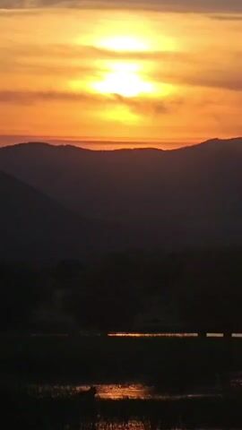 A video shows a sunset with the sun as a bright yellow orb in the center of the frame. The sun is surrounded by streaks of orange and yellow clouds. Below the sun are silhouetted mountains and trees. In the foreground is a body of water reflecting the colors of the sunset.