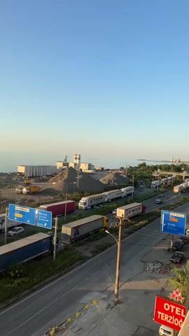 The video shows a highway with multiple lanes of traffic. There are many semi-trucks, some of them parked along the side of the road. In the background, there is a body of water, possibly a sea, and a clear blue sky. There are also some industrial buildings, piles of gravel, and construction equipment near the water. A sign for "OTEL TERZİOĞLU" is visible in the lower right corner of the frame. The time of day appears to be late afternoon or early evening, judging by the lighting.