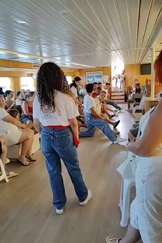 The video captures an indoor scene aboard a vessel, possibly a ferry or cruise ship, with a group of people engaged in an activity. The room is illuminated by natural light streaming in from windows and artificial lighting from overhead fixtures. The floor is wooden, and the ceiling is paneled. Several individuals are seated and standing, observing a performance or demonstration.

In the foreground, a woman with curly dark hair, wearing a white t-shirt and blue jeans, stands with her back to the camera. She has a red sash tied around her waist.

In the center of the frame, a man in a white t-shirt and blue jeans is kneeling on one knee, leaning forward with his arms extended. He appears to be interacting with another man who is also kneeling and looking at him. Behind them, several other men and women are seated or kneeling, watching the activity unfold. Some individuals in the background are standing near a staircase.

The atmosphere seems relaxed and engaging, with people dressed casually in t-shirts, shorts, and jeans, suggesting a daytime leisure activity. The overall impression is of a group participating in a recreational or educational session on a boat.