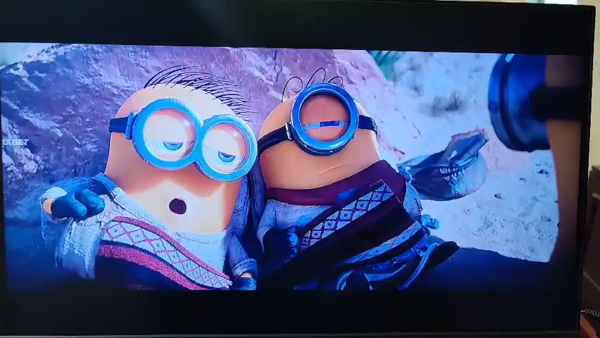 The video displays a scene from a movie featuring animated yellow characters, commonly known as Minions, in a desert-like setting. The characters are dressed in simple, light-colored tunics and wear large goggles. One Minion in the foreground holds a tall stick with a red ribbon tied to it. Several other Minions are visible around it, some appearing to be looking up at the one with the stick. The ground is sandy and uneven, with rocks and formations suggestive of a desert landscape. In the background, there are more rocky formations and a clear, light blue sky, indicating a bright day. The Minions appear to be either listening intently or awaiting instruction. There is a logo that says "1XBET" in the upper left corner of the screen. The overall mood seems neutral, with the Minions exhibiting characteristic expressions of curiosity or slight confusion. The landscape does not provide specific cues to pinpoint the location as Shahrisabz City, Uzbekistan.