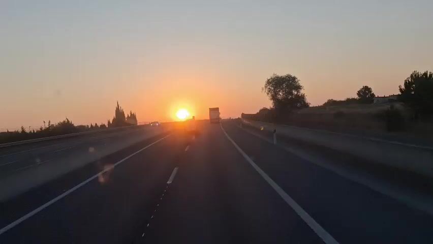 The video shows a point-of-view shot from a vehicle traveling on a highway at sunrise. The sun is a bright, orange orb low on the horizon, casting a warm glow over the scene. The highway has multiple lanes, with other vehicles, including cars and a truck, visible in the distance. The landscape on either side of the highway features sparse vegetation and some trees. The sky is a soft gradient of orange and pale yellow. The shot is steady, indicating the vehicle is moving at a consistent speed.