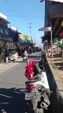 The video shows a street scene with several parked motorcycles. One motorcycle in the foreground is white and red with a license plate reading G 2437 YU. Indonesian flags are displayed on poles along the side of the road, which appears to be under an overpass. There are buildings and shops lining the opposite side of the street. People are visible walking and standing near the shops, and some are sitting on the side of the road. The sky is blue and clear, indicating it is daytime.