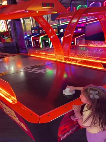 Two young girls are engaged in a game of air hockey within a vibrant arcade setting. The central focus is an orange and black air hockey table, brightly illuminated by glowing orange LED strips along its edges and overhead arches. The brand name "DYNAMO" is visible on the side of the table, and the word "AMMO" appears repeatedly on the translucent back panels. An overhead scoreboard displays "3 6."

On the right side of the table, a girl with long, dark brown hair, styled with a grey bow, stands facing away from the camera. She wears a yellow top and light-colored pants, holding a white air hockey paddle with both hands, poised for play. On the opposite side, partially visible, another girl with long brown hair and a yellow top holds her paddle, looking intently across the table, appearing engaged and focused on the game. A small, light-colored puck rests on the black playing surface.

The background reveals a bustling arcade environment, filled with the colorful glow of various gaming machines. A basketball arcade game, clearly labeled "NBA HOOPS" with a score of "1300" visible, stands behind the girl on the left. Other arcade cabinets with bright neon lights in shades of red, blue, green, and purple line the walls, creating a lively and energetic atmosphere. The scene is indoors, likely in the evening, given the reliance on artificial, colorful lighting typical of entertainment centers. The girls appear to be enjoying their game in this fun, competitive setting.