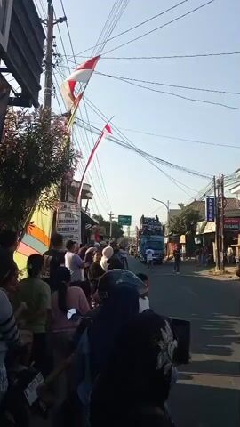 A group of people are gathered on the side of a street, some on motorcycles and others standing. There are Indonesian flags displayed, one large one attached to a pole and another smaller one further down the street. A blue truck with a large sound system on top is driving down the street, and the people appear to be watching it. The sky is clear and blue, suggesting it is daytime. There are power lines overhead, and buildings line the street.