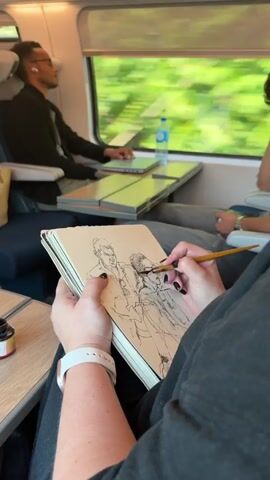 A person on a train is drawing in a sketchbook with a brush pen. The sketchbook is open and filled with a black ink sketch of two people. The person drawing has black nail polish and a white wristwatch. Next to the sketchbook is a small bottle of ink. In the background, a man with glasses and earbuds is sleeping, with a laptop on a tray table. A water bottle is also visible on the tray table. The train window shows a blurry green landscape, indicating the train is in motion.