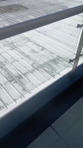 The video shows a mountain landscape from a rooftop perspective. The roof is made of gray corrugated metal sheets. A tall, white, multi-tiered cylinder stands on the roof. A gray railing runs along the edge of the roof. In the background, there's a scattering of houses among trees. Further back, two mountains rise against a clear blue sky. There's a small patch of white visible on the peak of the mountain on the left. The camera zooms in slightly on this mountain.
