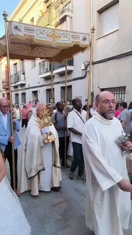 A religious procession is moving down a paved street. A priest in white vestments is at the center, carrying a golden monstrance. He is walking under a canopy supported by four poles held by men in front and behind him. The canopy is light-colored with gold trim and decorations, including a stylized sun and grapes. Many people are walking in the procession, including a young girl in a white dress at the front left, holding white flowers. Others in the procession are adults of various ages and ethnicities. Buildings line both sides of the street, featuring multiple stories and balconies. Some onlookers stand on the sidewalks. The weather appears sunny.