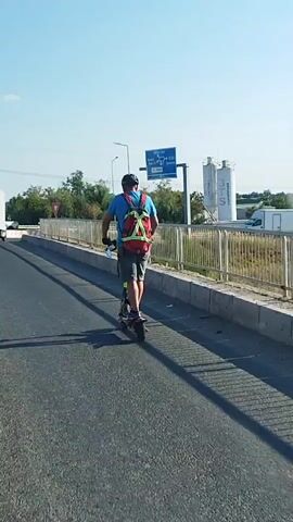 A man rides an electric scooter down a paved road. He is wearing a blue shirt, grey shorts, a black helmet, and a red backpack with a reflective green vest over it. The scooter has a black frame and red accents on the wheels. The road is bordered by a concrete barrier with a metal fence. In the background, there are trees, a large blue sign with white lettering, a white truck, a black car, and two large silos. The sky is clear and blue. The scene appears to be a sunny day.